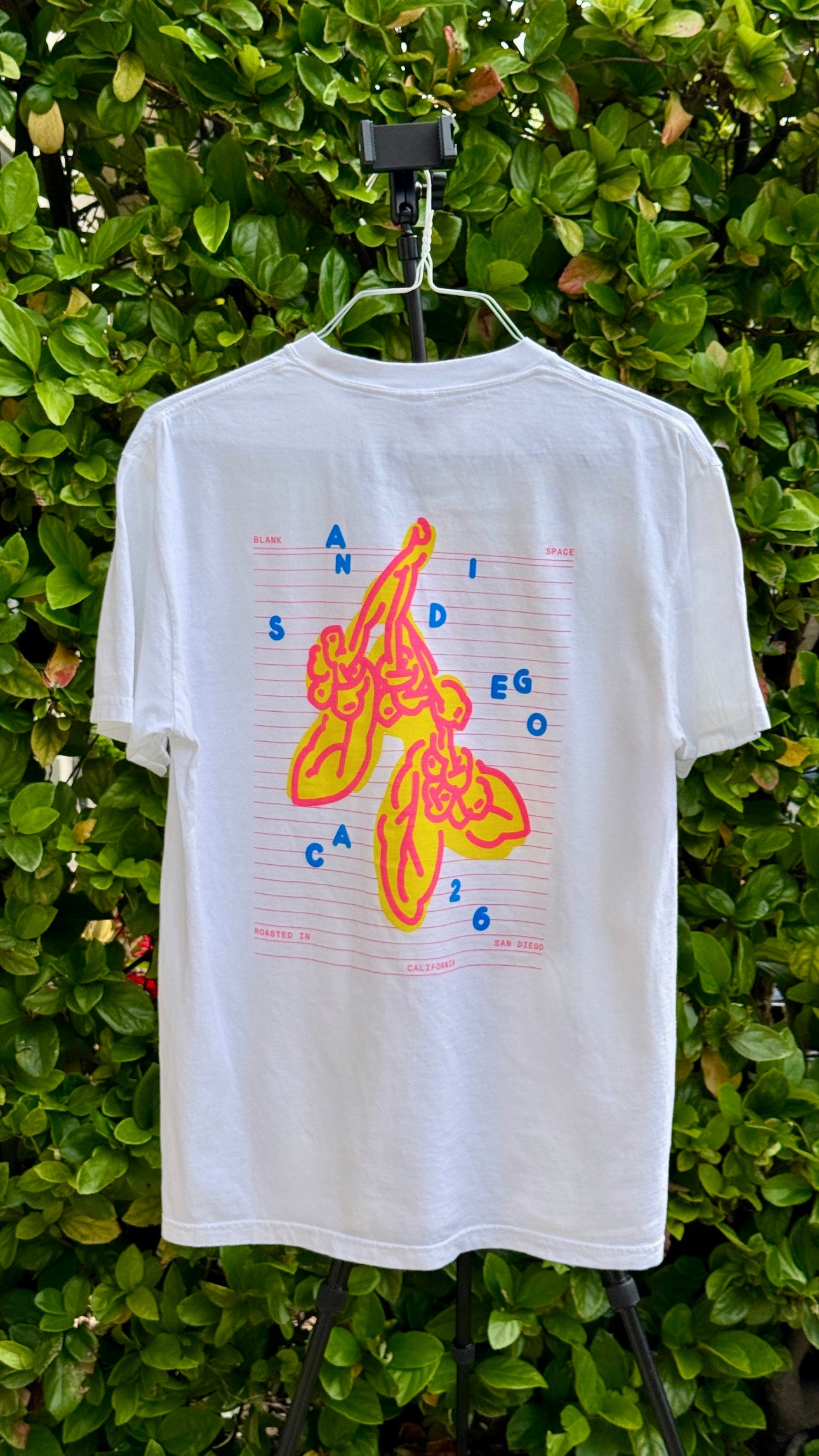 White t-shirt with colorful graphic design on a hanger against a green leafy background